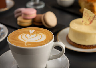 Café  con arte latte en un primer plano acompañado de distintos tipos de tortas con foco diferenciado.