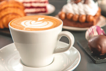 Café  con arte latte en un primer plano acompañado de distintos tipos de tortas con foco diferenciado.
