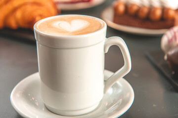 Café  con arte latte en un primer plano acompañado de distintos tipos de tortas con foco diferenciado.