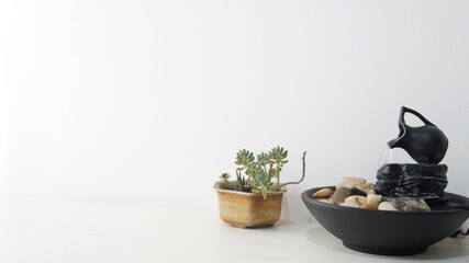  Zen water fountain and a small succulent plant placed on top of a white wooden table. A white bright wall as a background  