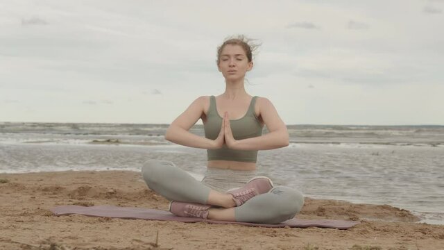 Slowmo shot of young concentrated Caucasian woman practicing yoga alone outdoors in morning, sitting in lotus pose with praying hands on yoga mat on sandy beach by water
