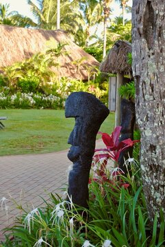 A stone tiki, white spider lilies, and a red Florica Cordyline plant a in Fiji garden. Tikis are large wooden or stone carvings or statues of human forms common throughout the Fiji islands.