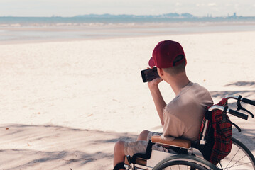 Happy disabled teenage boy on wheelchair use social media, Sea beach background, Lifestyle...