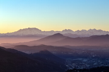 Naklejka na ściany i meble View of the misty and fog surrounding the mountains of the Italian Alps at winter sunset. Layers of mountains. Nature tourism.