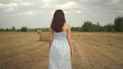 Fototapeta na wymiar A young woman walks in a field with haystacks. Girl with glasses and a blue dress. Back view.