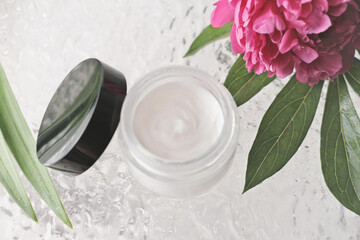 Obraz na płótnie Canvas white cosmetic face cream in a glass bottle with black lid. peony flower and shower door. self care and beauty treatment. top view