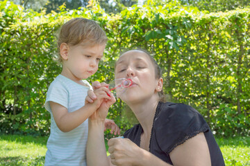 Cute little boy and his mother having fun blowing soap bubbles in the park. Funny outdoor activity for children in a beautiful summer sunny day
