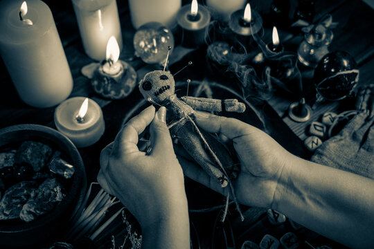 Sorceress or witch sticks needles into voodoo doll at ritual table with pentagram, burning candles and other occult objects. Voodoo witchcraft, spirituality and occultism concept, black and white