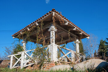 White wooden gazebo in the garden overgrown with plants on the blue sky background