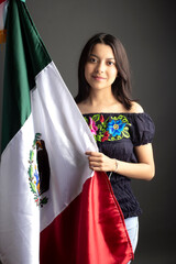 Beautiful Young hispanic woman with flag of Mexico wearing the typical costume in grey background. Independence day in Mexico. September celebration in Mexico.