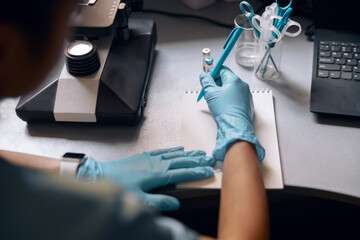 Woman in gloves writes in notebook near microscope at table in laboratory
