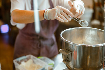Baker in gloves pours powder ingredient into bowl making dough in workshop
