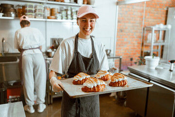 Lady in cap holds tray of croissants decorated with burnt cream in bakery