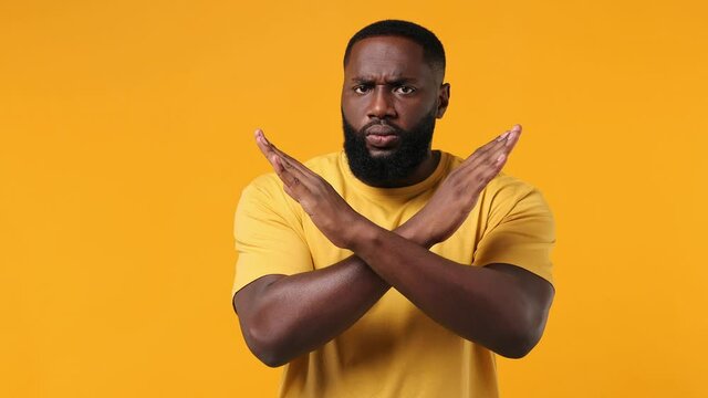 Serious strict severe young bearded african american man 20s years old wears orange t-shirt say no hold palm folded crossed hands in stop gesture isolated on plain yellow background studio portrait
