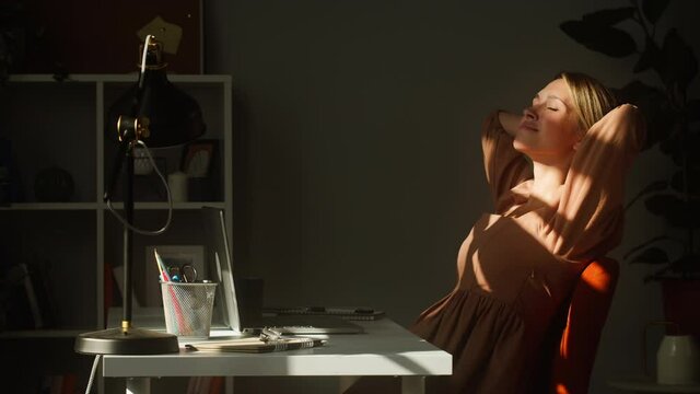 Young smiling woman resting at workplace. Happy business woman lying on chair and relaxing. Remote workplace, home office concept. Executive female manager sitting in sun rays. 