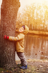 little happy four year old kid boy, who squeezed his eyes shut, in fall clothes hugs a tree trunk during a walk in the autumn park near the pond. flare
