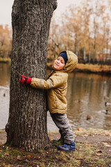 little happy four year old kid boy, who squeezed his eyes shut, in fall clothes hugs a tree trunk during a walk in the autumn park near the pond
