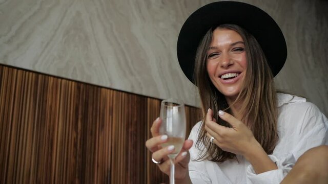 cheerful girl in a black hat laughs and drinks champagne sitting on the bed at her hotel. High quality FullHD footage
