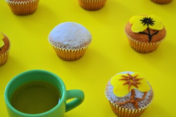 Rows of muffins decorated with powdered sugar and yellow flower shaped icing on a yellow background next to a cup of tea
