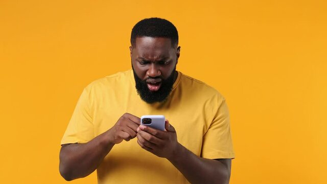 Sad irritated young african american man 20s wears orange t-shirt using mobile cell phone swear hears fake news unexpected rumor has some problems isolated on plain yellow background studio portrait