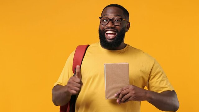 Excited young african american man 20s wears orange t-shirt backpack glasses hold book with blank screen workspace area show thumb up like gesture isolated on plain yellow background studio portrait