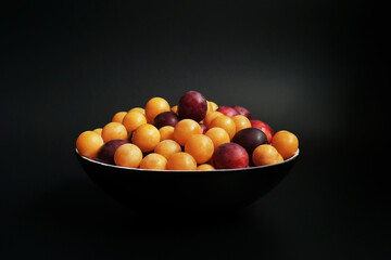 Ripe cherry plums of different varieties in a black bowl on the table. Black background 