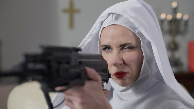 Close-up of serious woman in nun costume aiming with gun indoors. Portrait of beautiful Caucasian provocative passionate lady holding weapon looking away