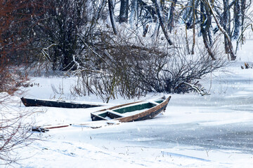 Snow-covered boat on the river in winter during a snowfall. Snow-covered trees by the river with a boat