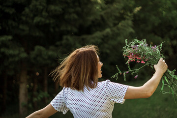 A girl in a white light dress is holding a bouquet of wild flowers over her head and spinning. Copy space, selective focus