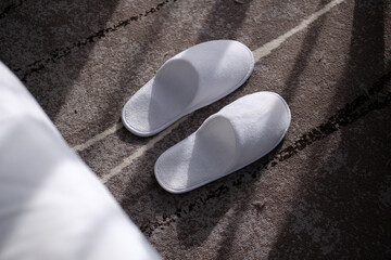 White cozy slippers on a grey carpet in a hotel room with window light shining through.