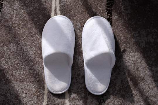 White cozy slippers on a grey carpet in a hotel room with window light shining through.