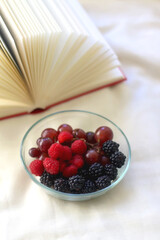 Bowl of raspberries, blackberries and grapes with open book. Selective focus.