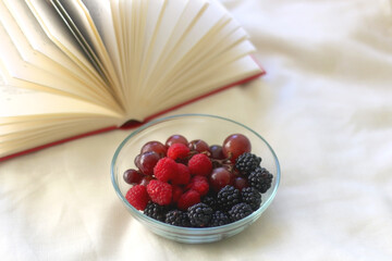 Bowl of raspberries, blackberries and grapes with open book. Selective focus.