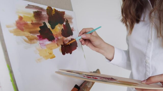 Close-up of wooman hands drawing picture with paintbrush and palette with colorful paints on easel indoors.