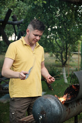 Naklejka na ściany i meble Young man putting firewood in mangal. Handsome athlete male builds a fire in a metal barbecue in the courtyard.