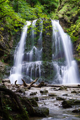 Ein imposanter Wasserfall mitten im Wald. Das Wasser fließt in einen Fluss. Es gibt Bäume rund um den Wasserfall.