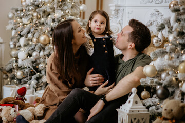 Young happy family dad, mom and daughter enjoy New Year and Christmas holidays. Father, mother and daughter are sitting on the background of a stylish decorated Christmas tree with toys and smiling