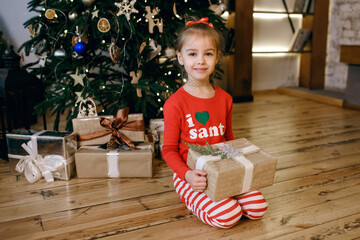 A little girl in pajamas sits near a decorated Christmas tree and holds a Christmas present in her hands. The atmosphere of a holiday and a happy childhood. Traditional New Year and Christmas holidays