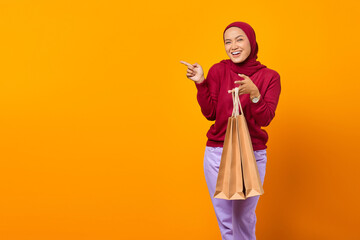 Cheerful Asian woman pointing to empty space and holding shopping bags over yellow background