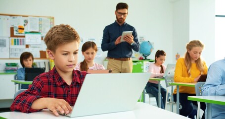Close up portrait of cute Caucasian teen boy pupil typing on laptop browsing online studying at computer science class, elementary school lesson. Man teacher helping kids using tablet on background