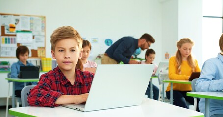 Cheerful little Caucasian kid schoolboy tapping on laptop surfing internet at computer science class, smiling at camera. Male teacher helping pupils on background. elementary school, Close up portrait