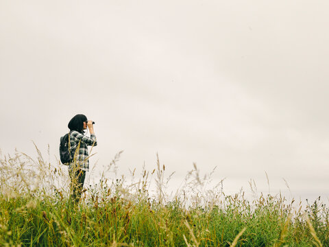 UK, Female hiker standing in grassy field, looking through binoculars