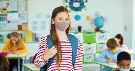 Portrait of happy Caucasian little girl wearing face mask stands in classroom and smiling to camera indoors Studying after coronavirus pandemic multi-ethnic primary school pupils learning during covid