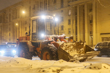 Fototapeta na wymiar Snow plow tractor with a large bucket drives along a city street during a blizzard