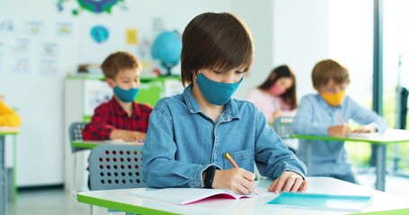 Caucasian little bright schoolboy in mask sitting at desk in classroom writing down at lesson and raising hand wants to answer question.