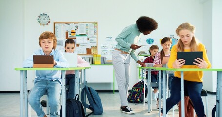 African American young pretty female teacher with gadget teaching pupils at school. Caucasian junior students boys and girls learning at school computer class using tablets and laptops devices