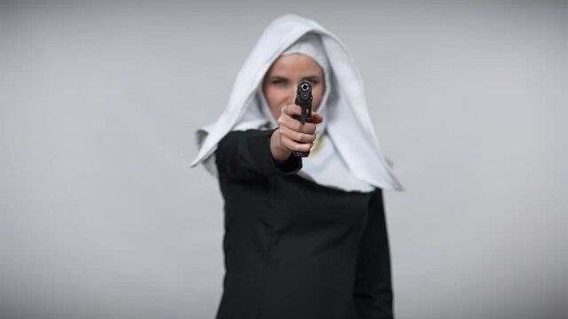 Rack focus from smiling woman in nun costume to handgun stretched at camera and back to face blowing at gun. Confident smiling gorgeous Caucasian lady posing with weapon at grey background