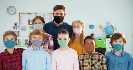 Portrait of handsome young Caucasian man teacher with diverse multi-ethnic pupils boys and girls wearing masks on faces standing in classroom looking at camera in good mood Elementary school, covid-19