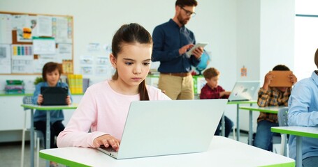 Close up of concentrated Caucasian little girl sitting at desk at school class typing on laptop Children studying in classroom using digital gadgets, computer science class modern elementary education
