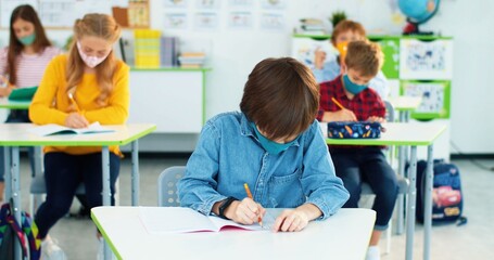 Portrait of Caucasian little bright schoolboy in mask sitting at desk in classroom writing down at lesson and raising hand wants to answer question.